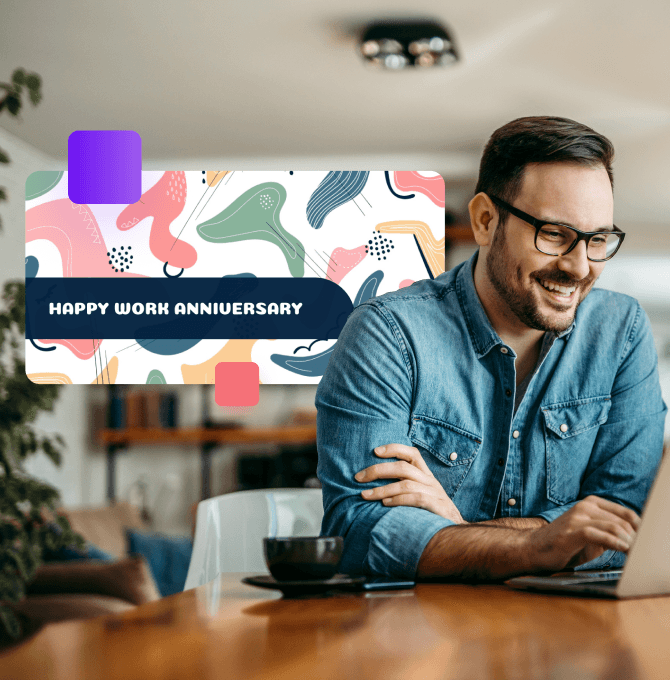 A guy with glasses in a blue shirt sitting in front of a laptop, smiling with a cup of coffee next to him.