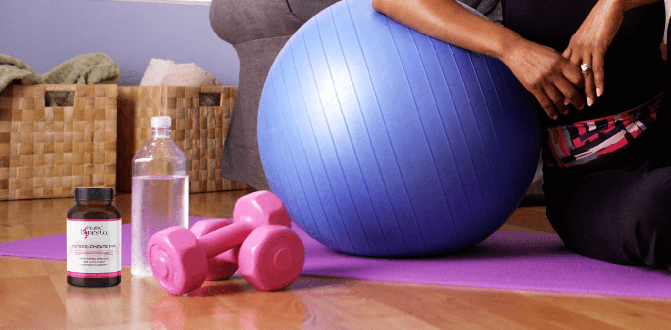 A woman next to a yoga ball, with a water bottle, two pink dumbbells, and the Healthy Bones Co. product.