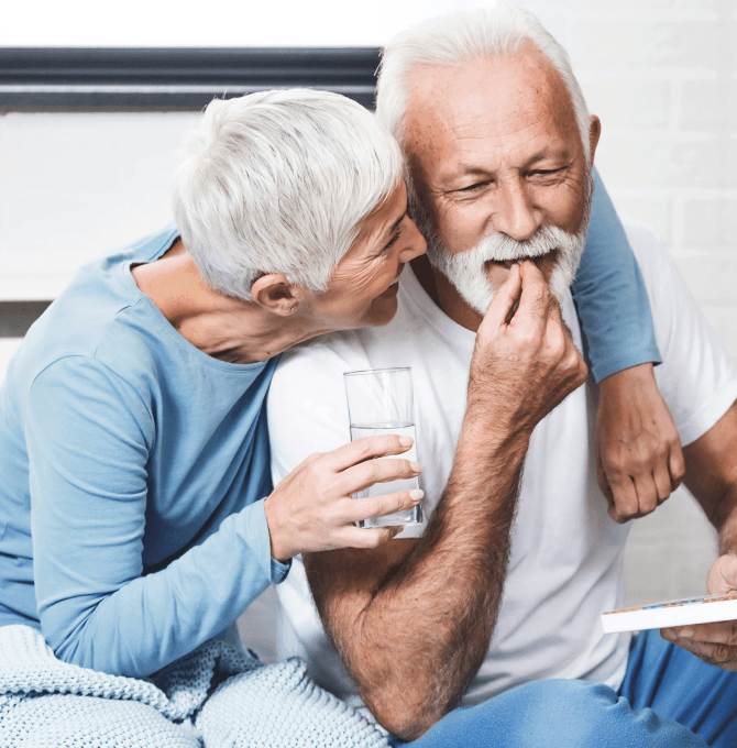 An older couple sits closely together, the man taking a pill and the woman holding a glass of water.
