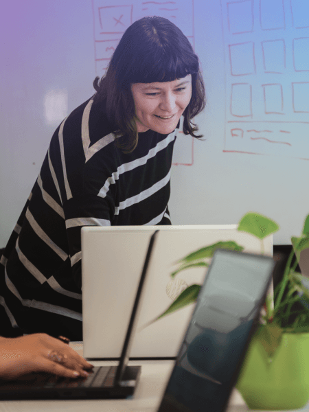 A woman smiling while working on a laptop, with a whiteboard in the background