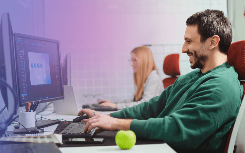 A man working on a computer with an apple in front of him, and a girl also working on a computer in the background